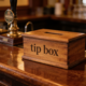 Pub tezgahında ahşap 'tip box' (bahşiş kutusu), arkada bira muslukları ve bardaklar