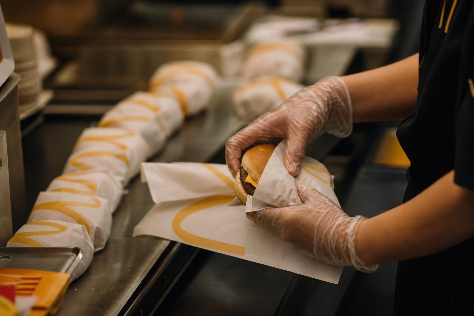 Fast food işçisinin eldivenli elleri hamburger paketlerken, küresel emek piyasasının sembolü