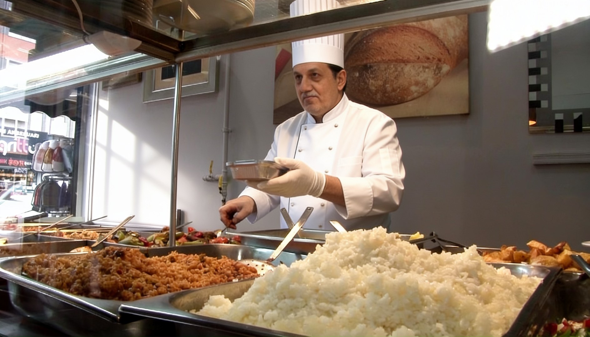 Bir Türk lokantasında beyaz önlüklü, şef şapkalı bir şef elinde yemek tabağıyla mutfak tezgahında duruyor, önünde metal tepsilerde pilav bulgur, kıymalı yemek ve salata, geleneksel lokanta atmosferi.